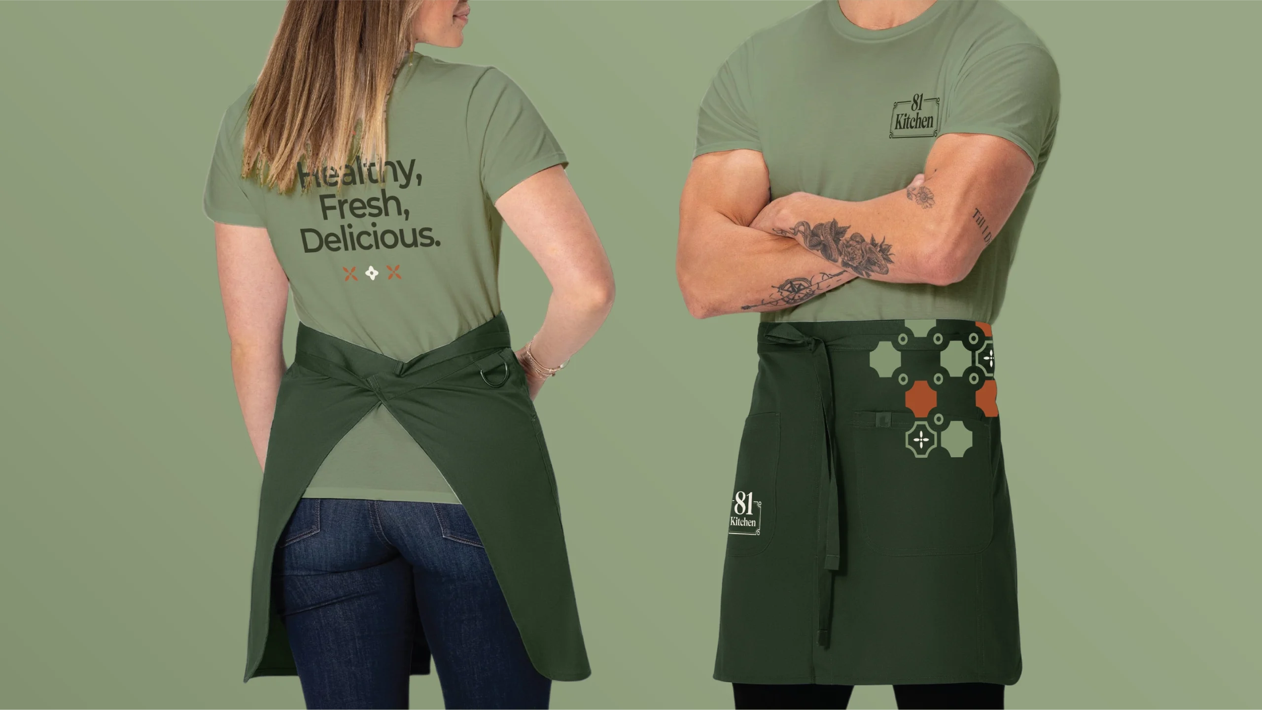 Two people in olive shirts and dark green aprons stand against a green backdrop; back view shows the slogan 'Healthy, Fresh, Delicious.'